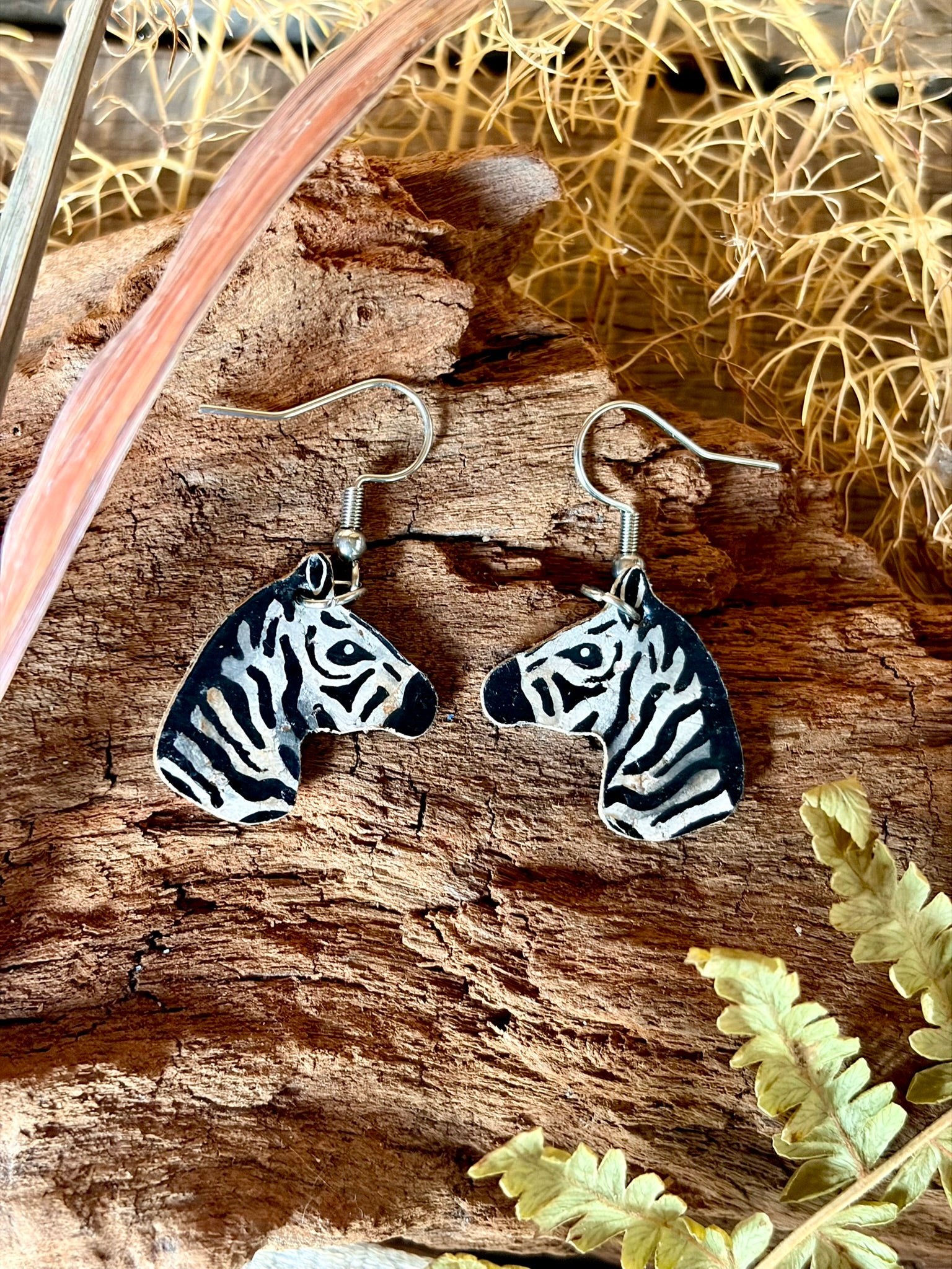 A pair of zebra-head dangle earrings with black and white striped detailings and silver fishhook wires, displayed on a textured piece of wood with dried botanicals.