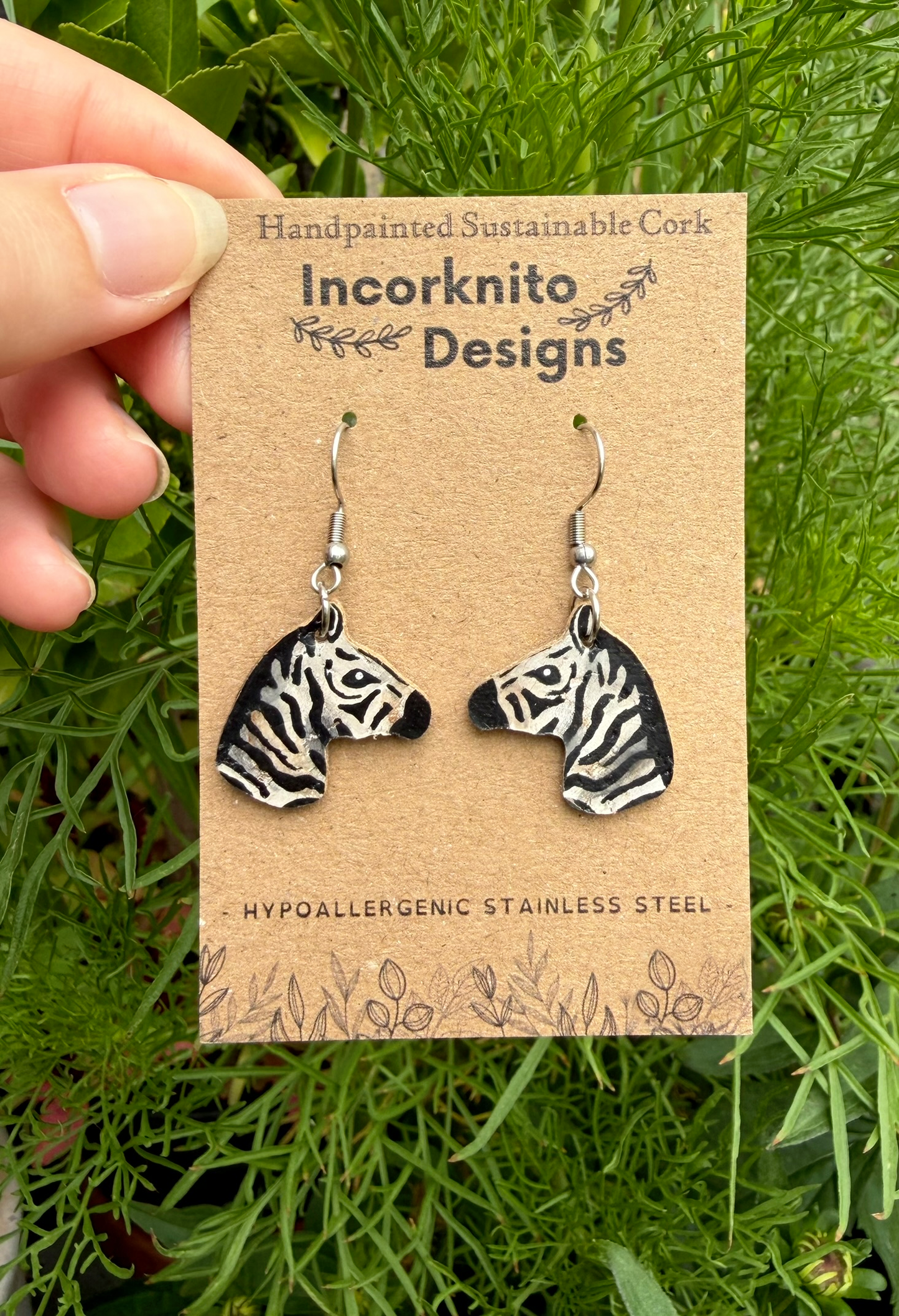 A hand holds a brown kraft display card featuring two zebra-head earrings on silver hooks. The card reads 'Incorknito Designs' and 'Handpainted Sustainable Cork' against a background of green foliage.