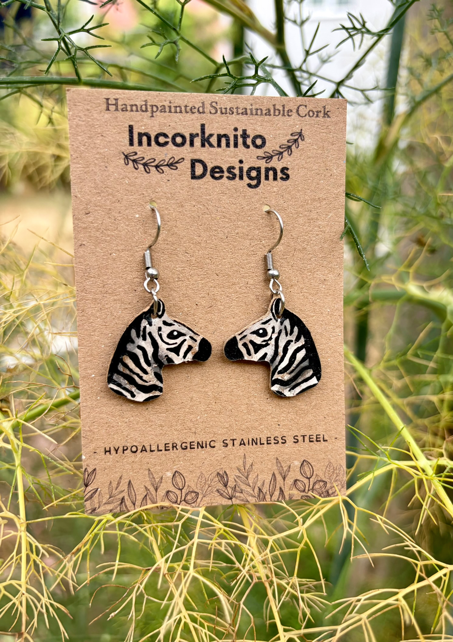 Handmade zebra head dangle earrings on a brown display card by Incorknito Designs, photographed outdoors among green and yellow foliage.