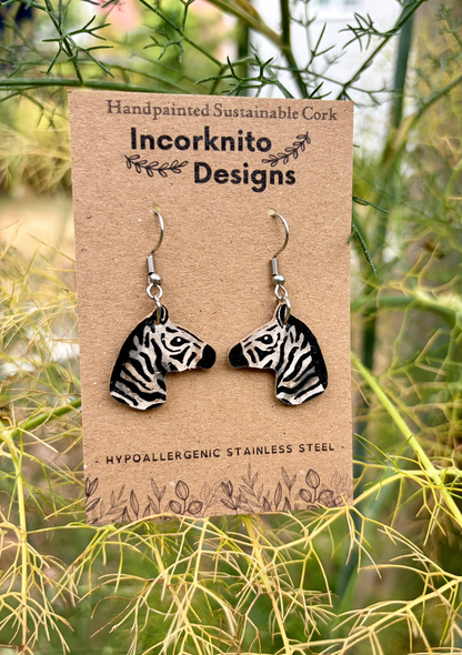 Handmade zebra head dangle earrings on a brown display card by Incorknito Designs, photographed outdoors among green and yellow foliage.