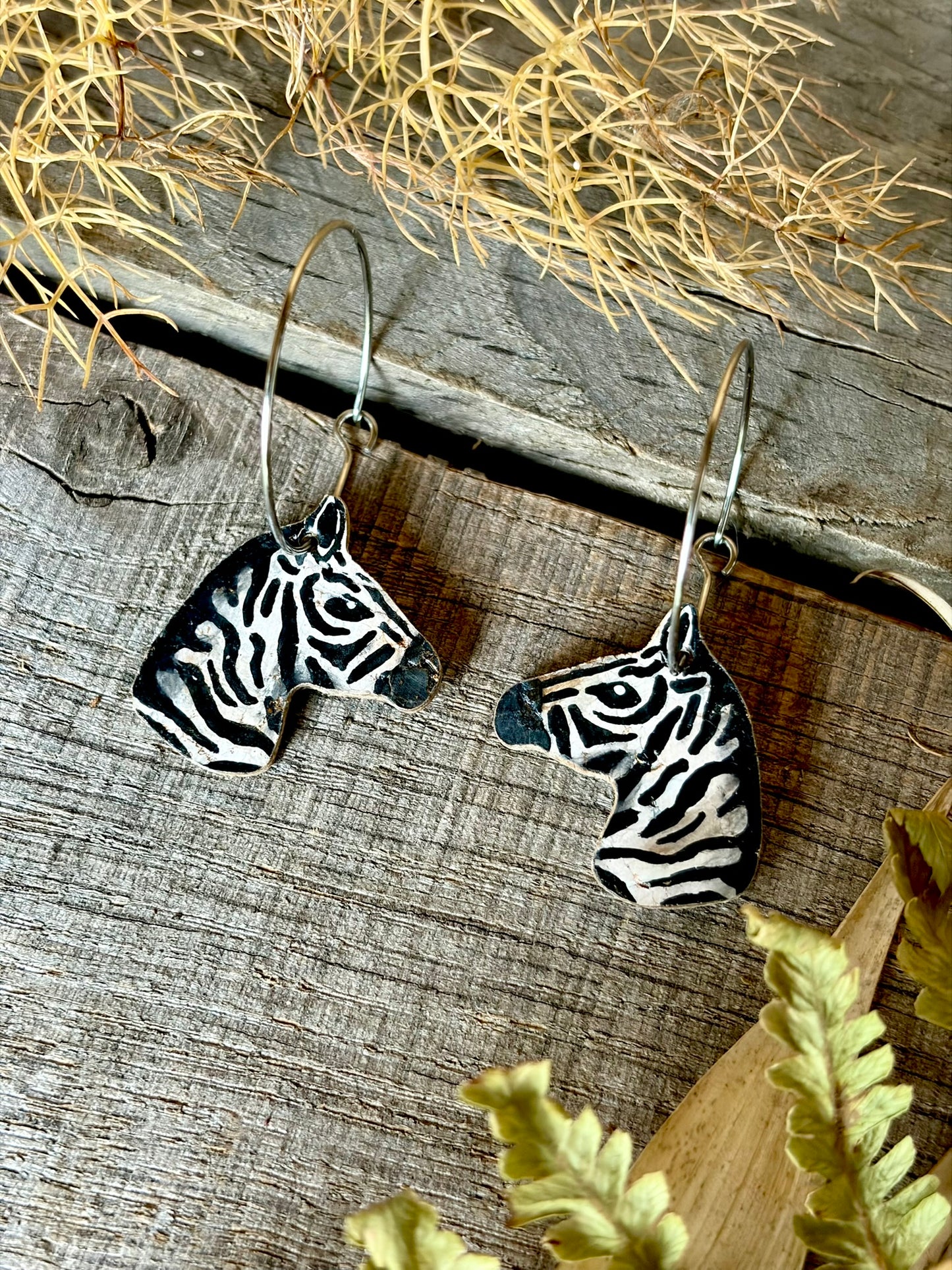 Zebra-patterned horse head drop earrings with silver-toned hooks, displayed on a rustic wooden surface with dried grass and green leaves.