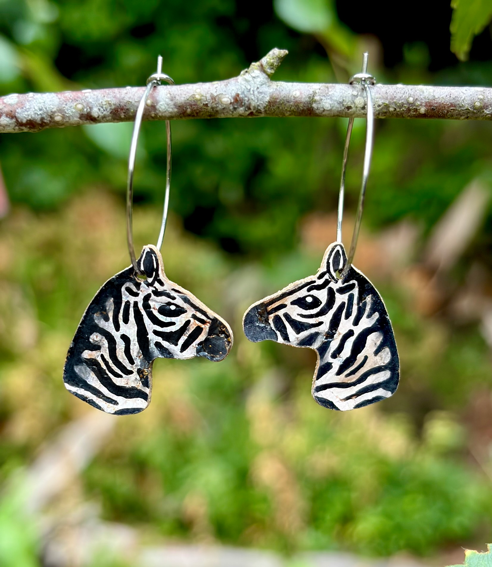 Pair of up close zebra-patterned drop earrings with stainless steel hoops, hanging from a tree branch against a blurred green outdoor background.