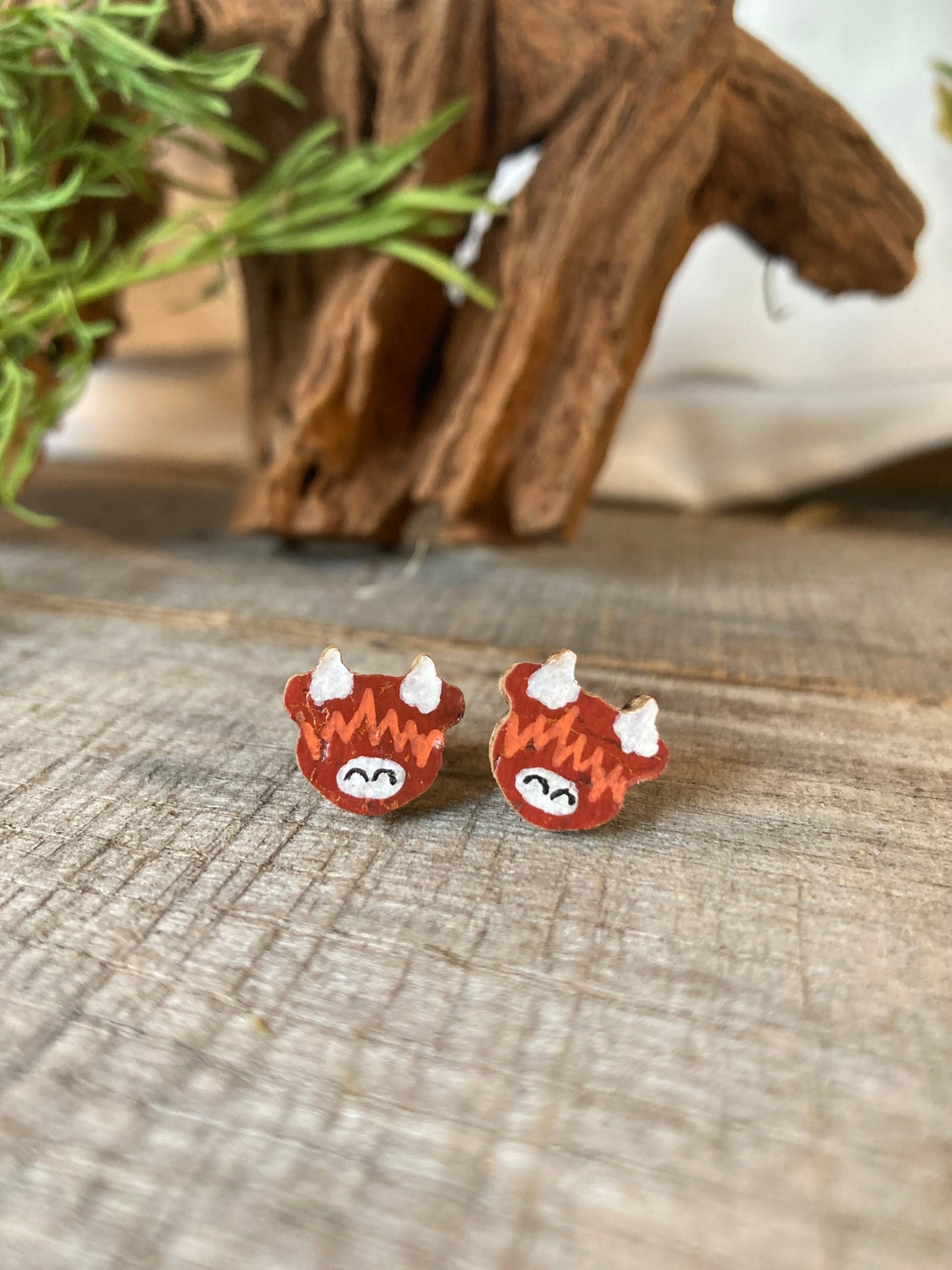 Handpainted highland cow cork stud earrings on a weathered wooden surface with driftwood and greenery