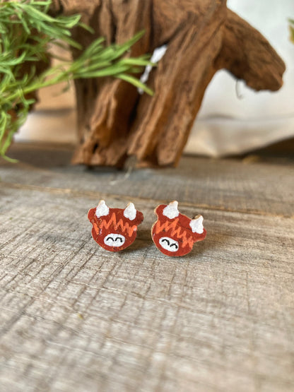 Handpainted highland cow cork stud earrings on a weathered wooden surface with driftwood and greenery