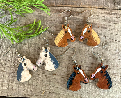 Set of horse cork earrings, three colourways - grey/white, bay/brown and chestnut/tan on wooden surface with natural greenery — handpainted sustainable wildlife jewellery