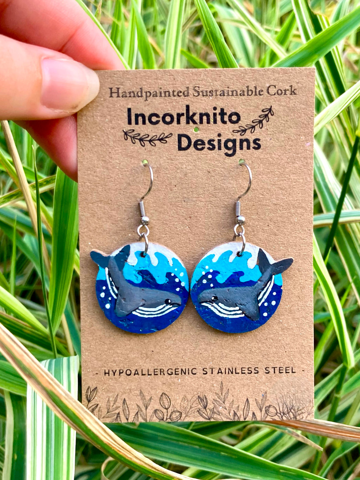 Handpainted humpback whale cork earrings on Incorknito Designs card with a whale swimming through vivid blue waves, held against lush green grass — handmade in Suffolk UK