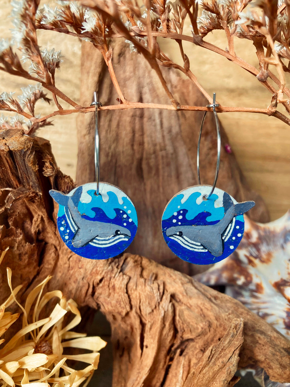 Humpback whale cork earrings on silver hoops hanging from a dried russet botanical branch against driftwood — handpainted sustainable cork jewellery