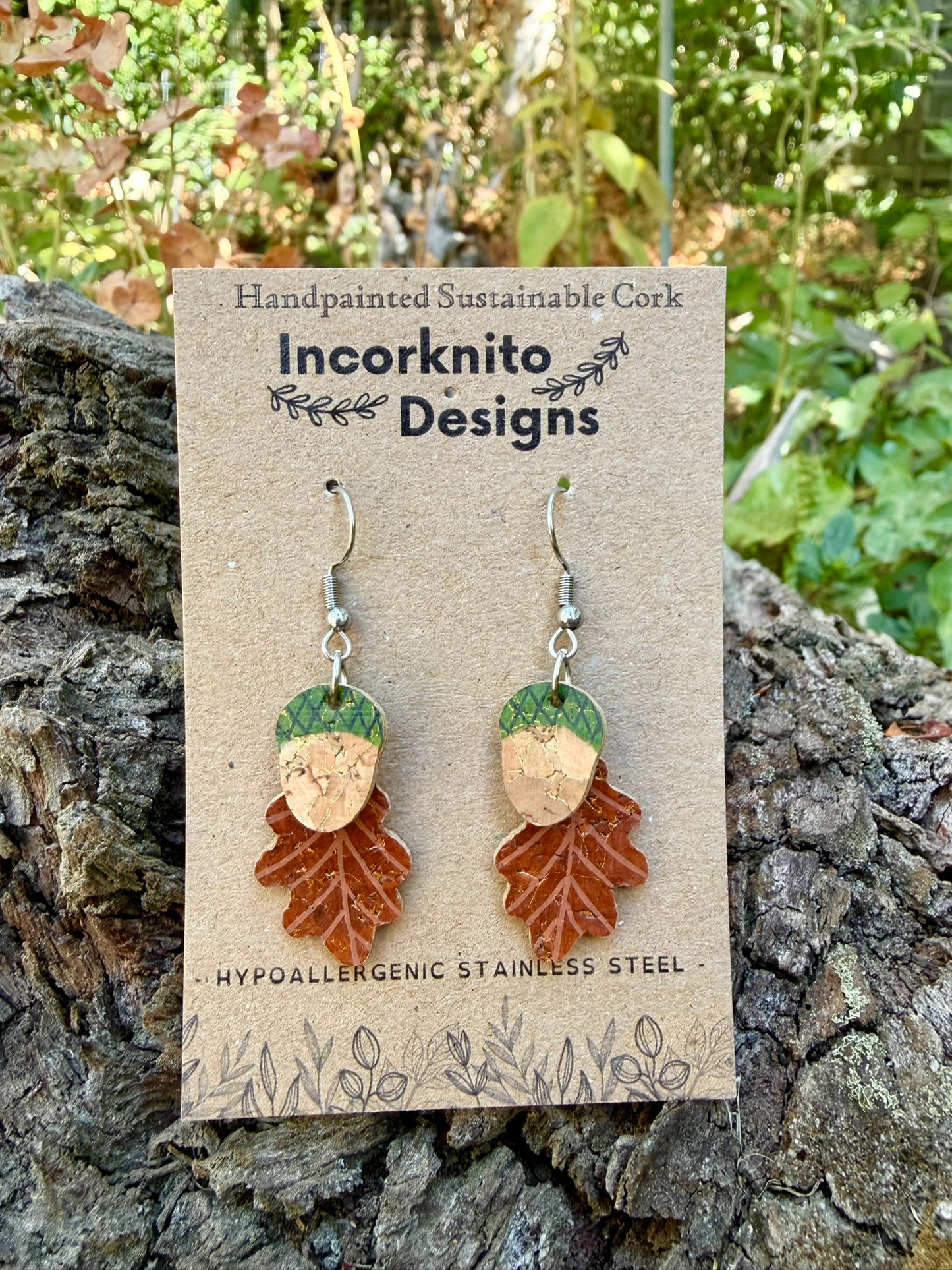 Acorn and oak leaf cork earrings in green colourway on Incorknito Designs card, propped against textured cork bark with autumn foliage