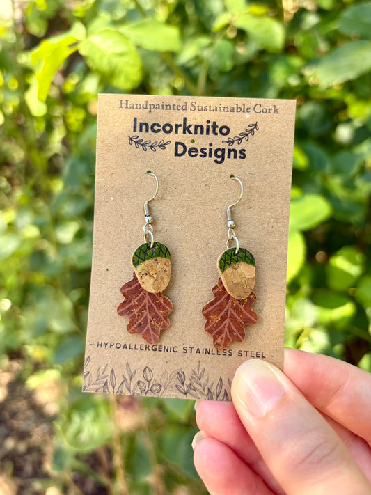 Handpainted cork acorn and oak leaf dangle earrings in green colourway, held on Incorknito Designs card against lush garden foliage