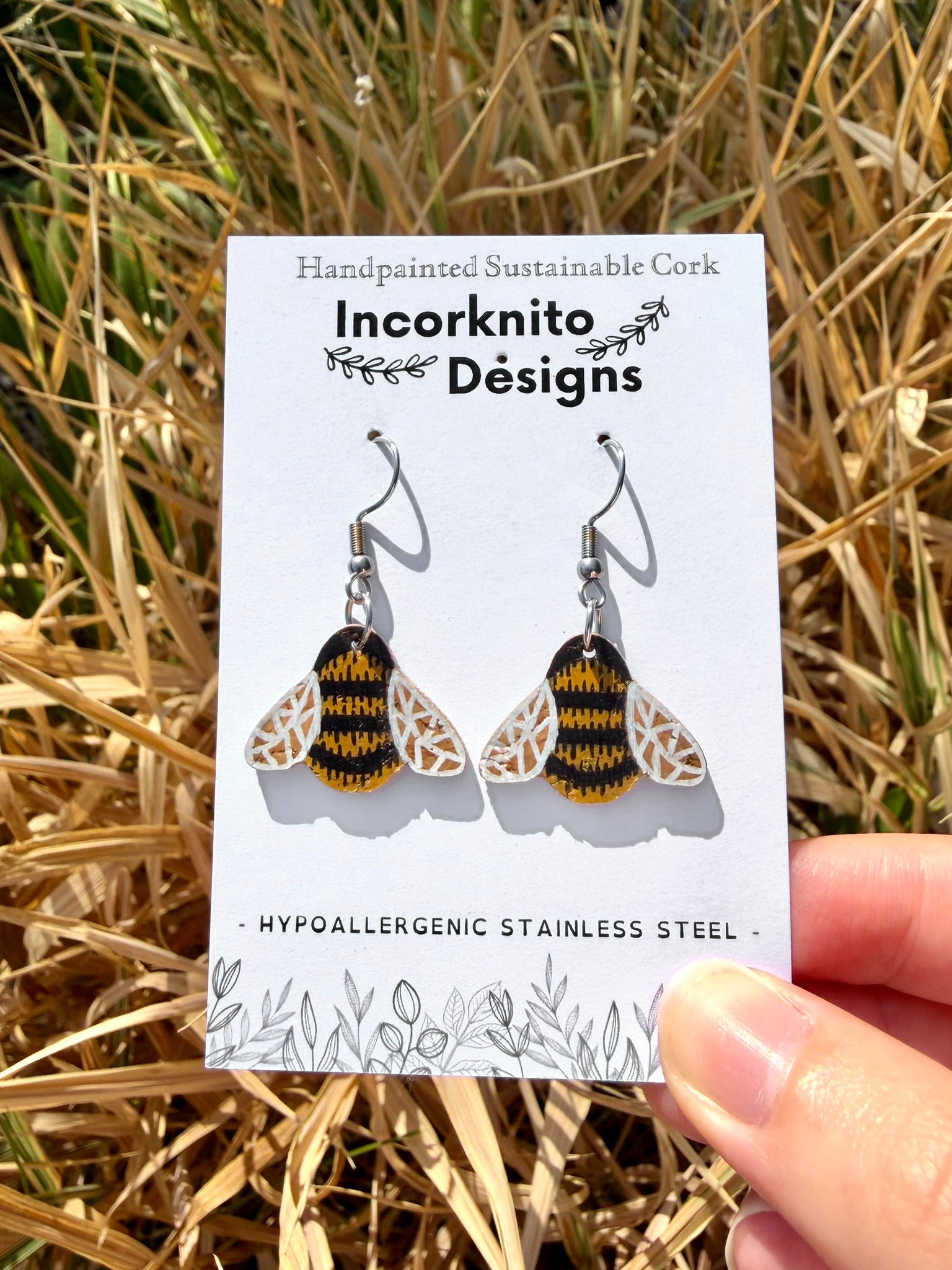 Cork bumblebee earrings with silver hook fastening on Incorknito Designs card, held against a golden dried grass field