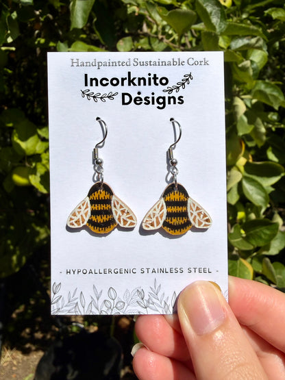 Cork bumblebee earrings with silver hook fastening on Incorknito Designs card, held against a green hedge