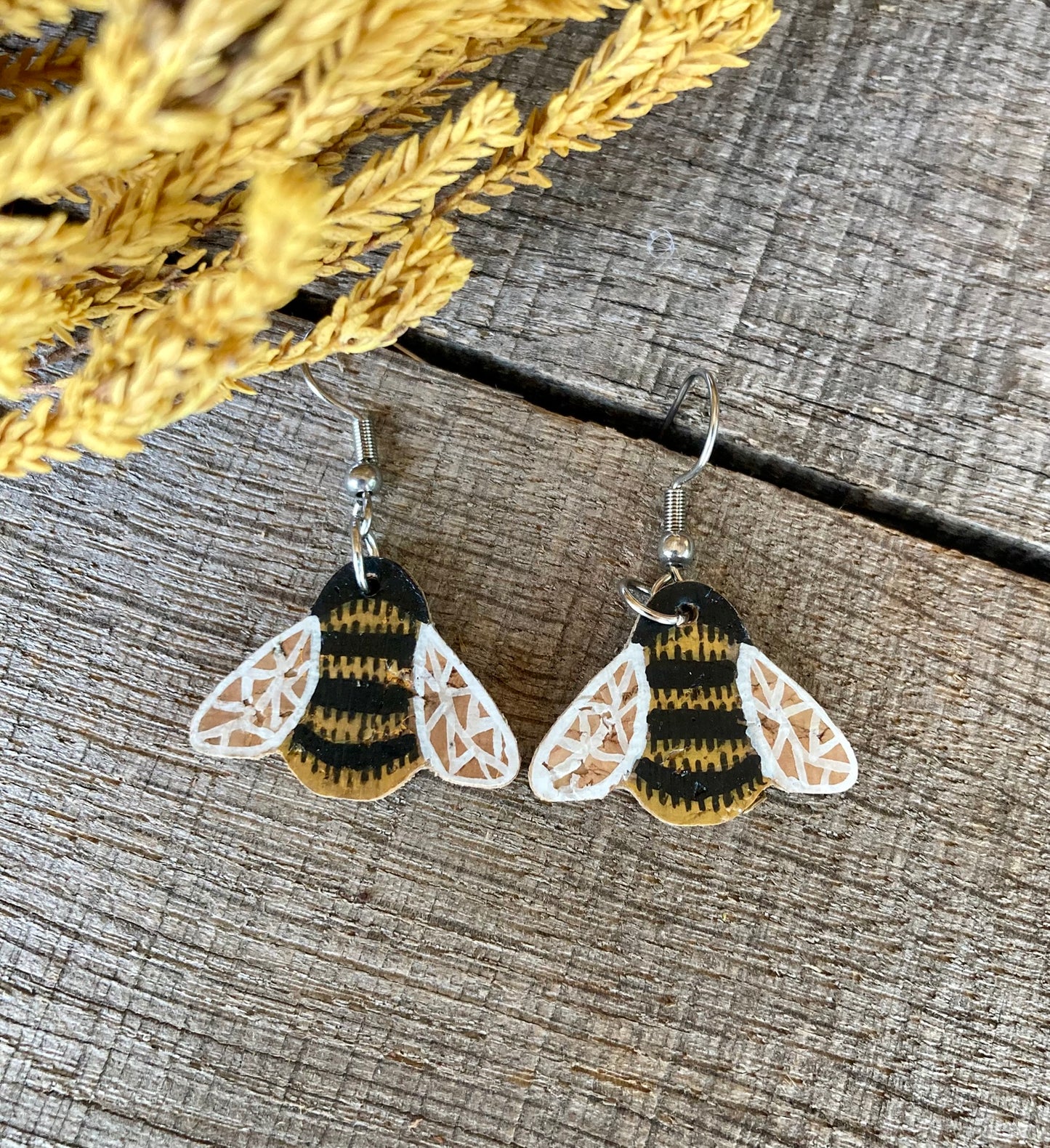 Handpainted cork bumblebee earrings with silver hook fastening, flat lay on weathered wood with golden dried grass