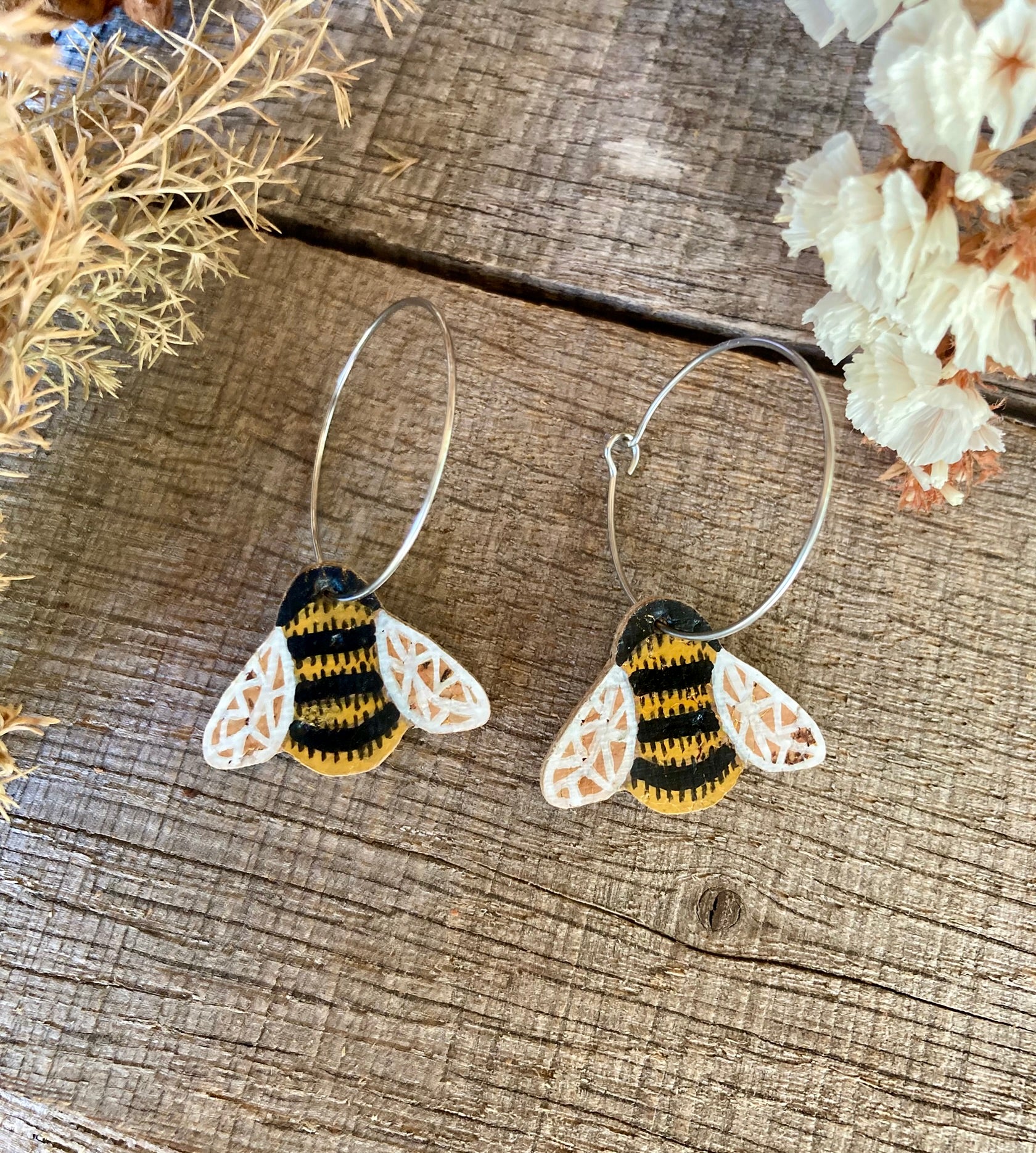 Handpainted cork bumblebee earrings on silver hoops, flat lay on weathered wood with dried pampas grass and white dried flowers