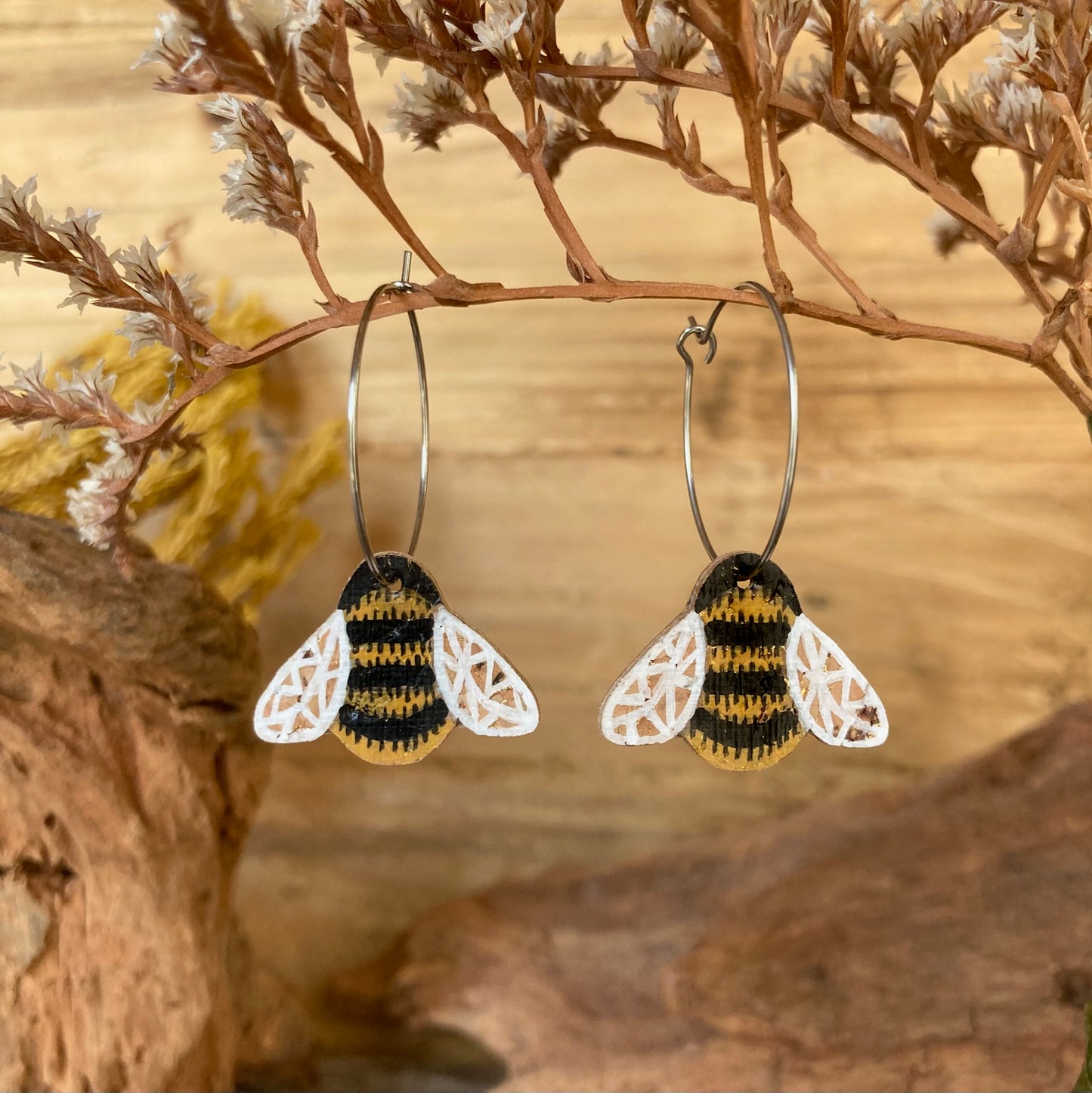 Cork bumblebee earrings on silver hoops, hanging from a dried branch with cork bark and dried flowers in warm tones