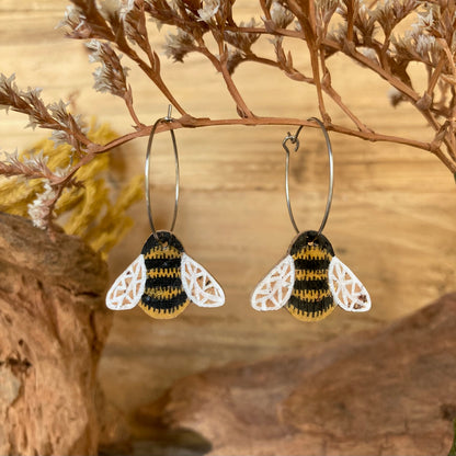 Cork bumblebee earrings on silver hoops, hanging from a dried branch with cork bark and dried flowers in warm tones