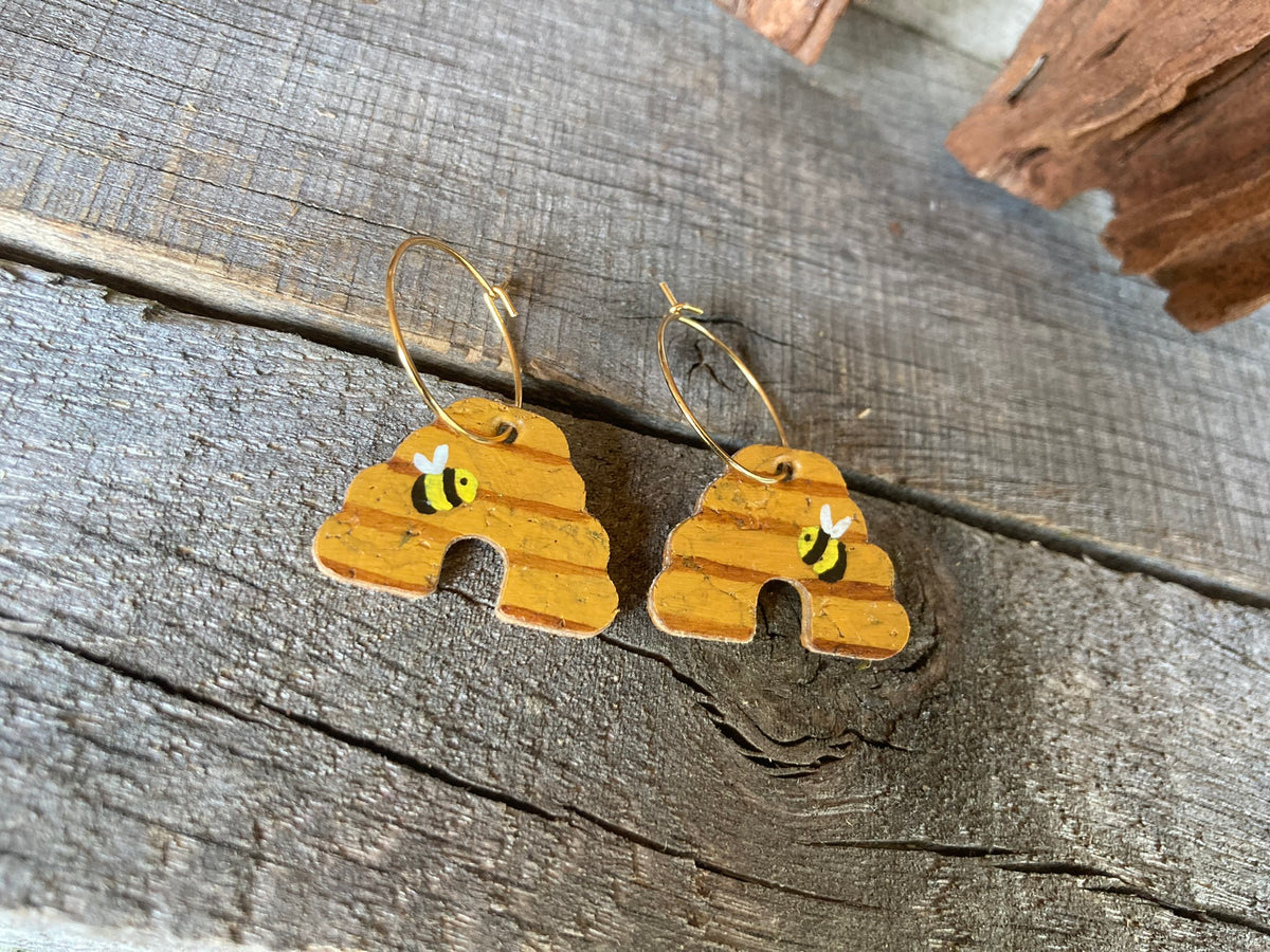 Cork beehive earrings on gold hoops, flat lay on weathered wood with cork bark — showing warm amber tones and bumblebee detail