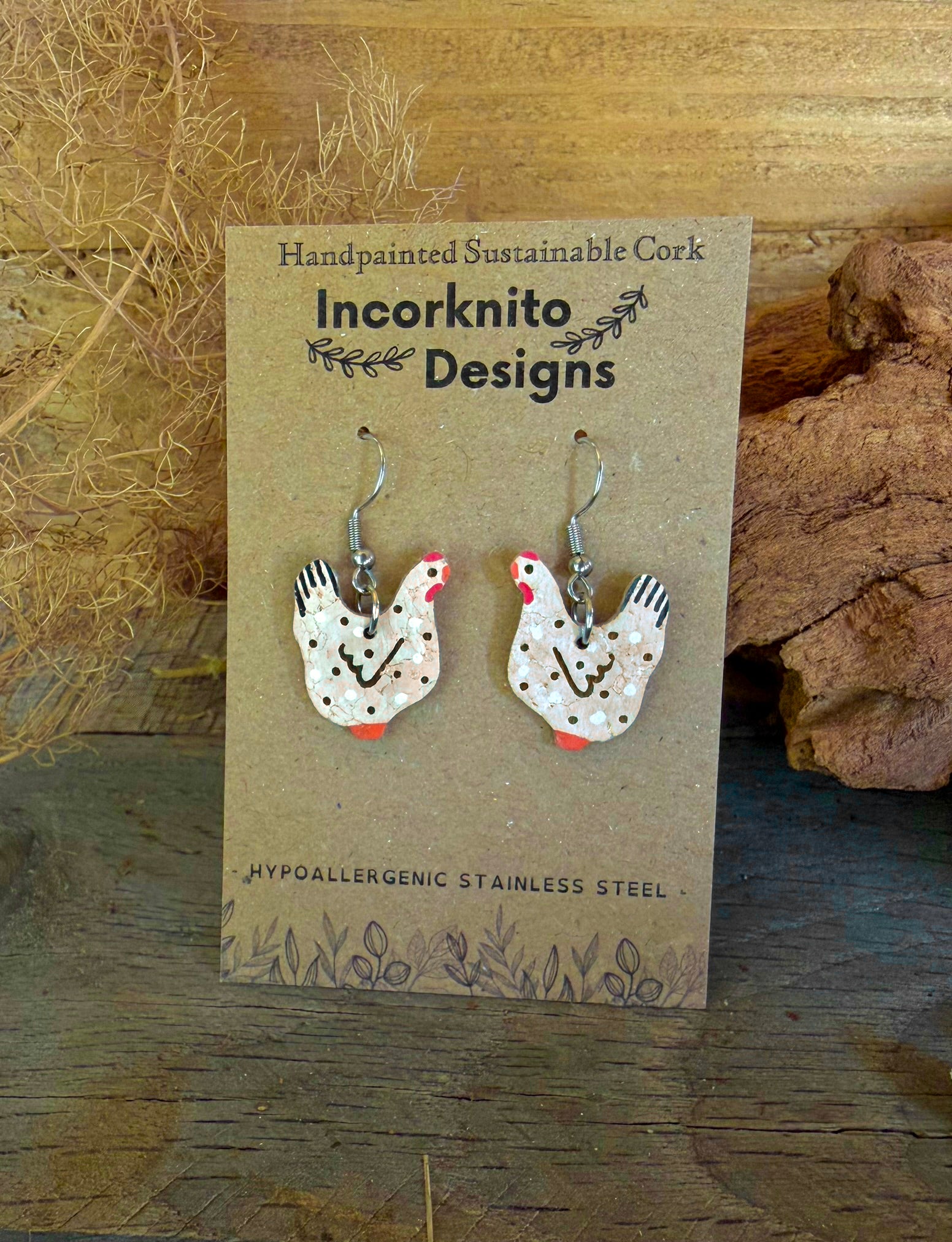 Handpainted black and white cork chicken earrings on silver hooks, displayed on Incorknito Designs branded card with dried grass and cork bark background