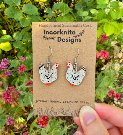 Handpainted black and white cork chicken earrings on silver hooks, displayed on Incorknito Designs branded card held in hand against garden flowers and greenery