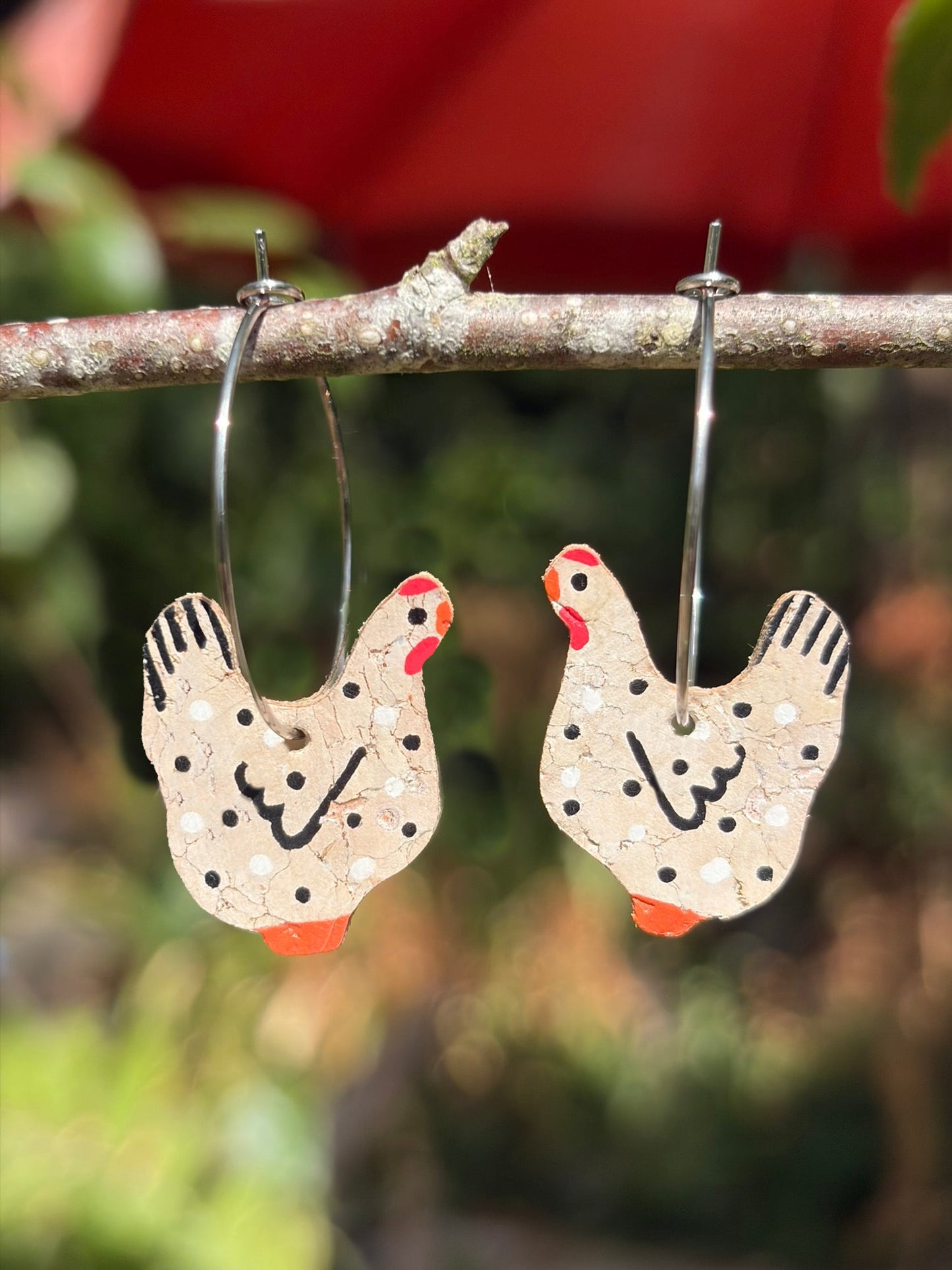Cork black and white chicken earrings on silver hoops, hanging from a garden branch with soft red and green bokeh background