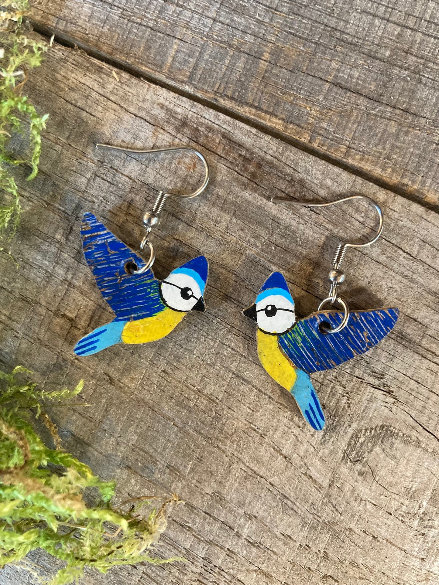 Blue tit cork hook earrings, handpainted, flat lay on weathered wood with moss
