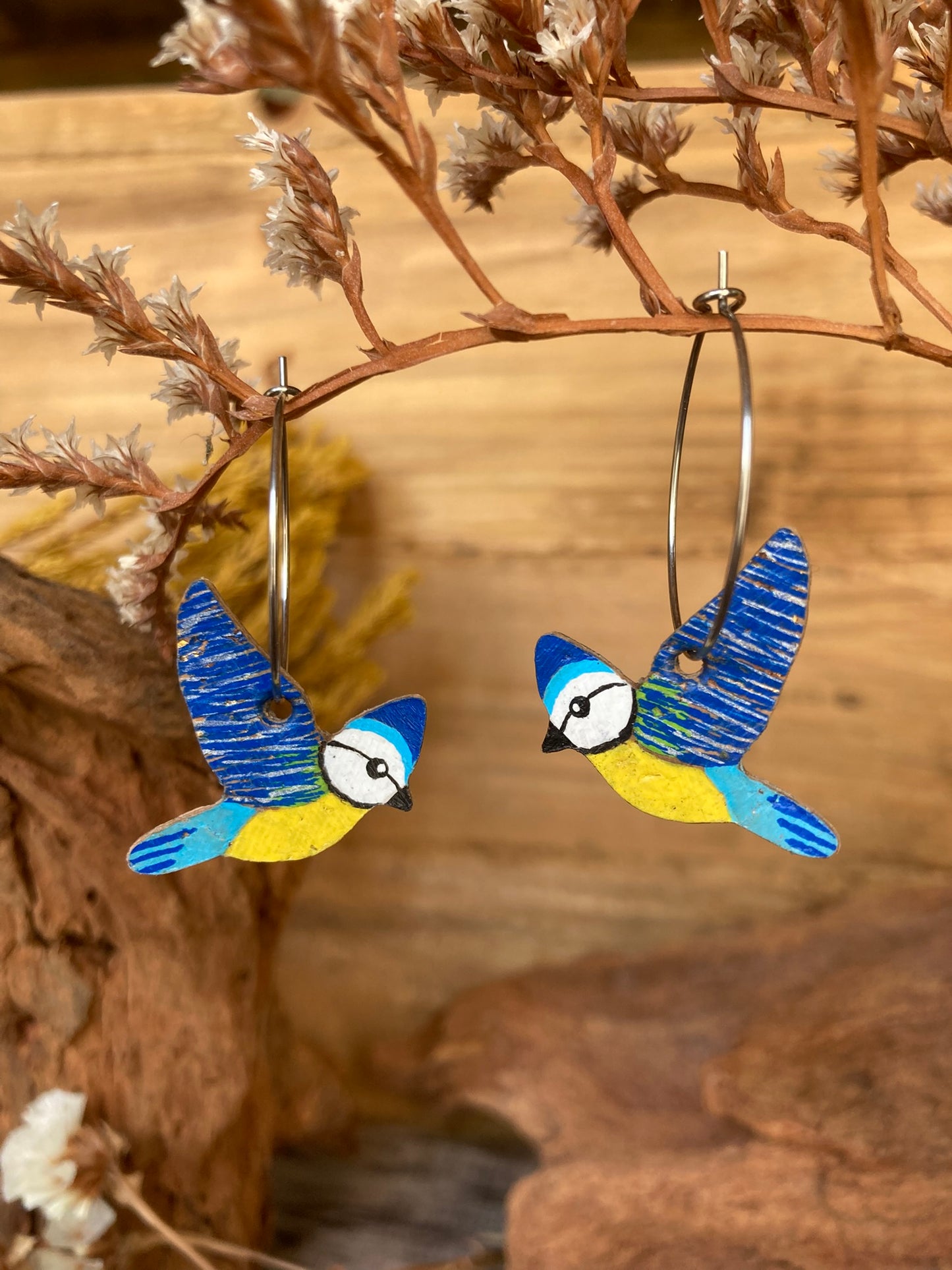 Blue tit cork hoop earrings, handpainted, hanging from a dried botanical branch