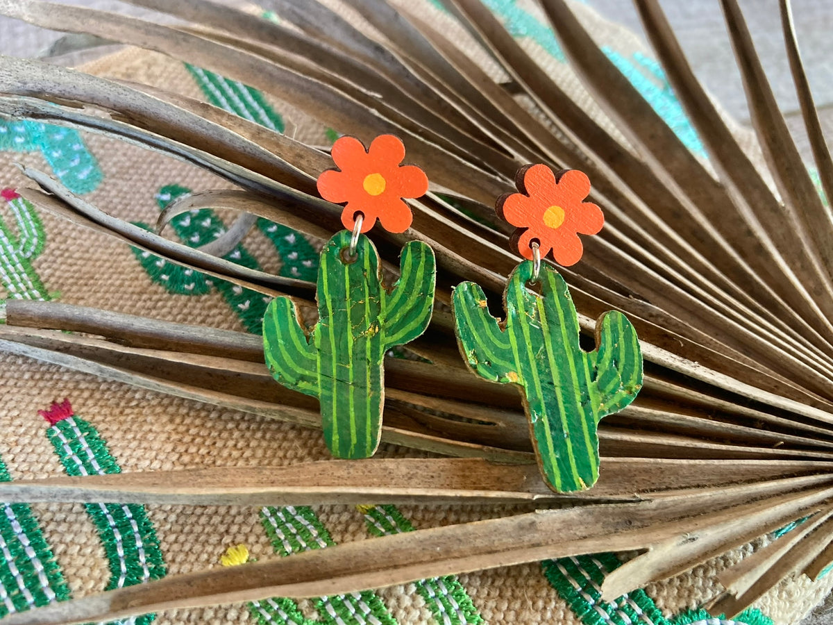 Handpainted cork cacti flower earrings in orange, displayed on a dried palm leaf with cactus fabric in the background