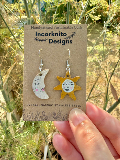 Handpainted cork sun and moon mismatch earrings on silver hooks, displayed on Incorknito Designs branded card held in hand against garden fennel and wildflowers