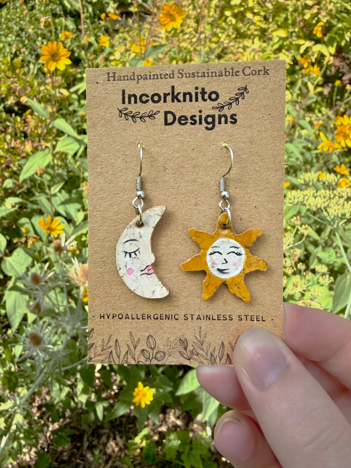 Handpainted cork sun and moon mismatch earrings on silver hooks, displayed on Incorknito Designs branded card held in hand against yellow garden flowers