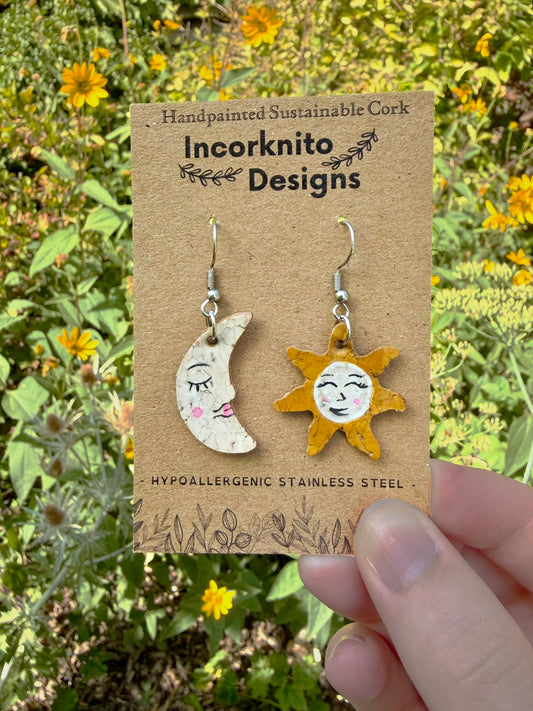 Handpainted cork sun and moon mismatch earrings on silver hooks, displayed on Incorknito Designs branded card held in hand against yellow garden flowers