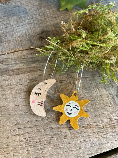 Handpainted cork sun and moon mismatch earrings on silver hoops, flat lay on weathered wood with moss and greenery background