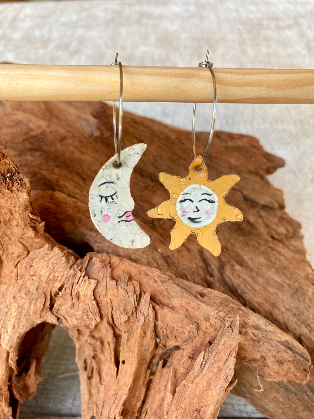 Handpainted cork sun and moon mismatch earrings on silver hoops, hanging from a wooden dowel against cork bark background