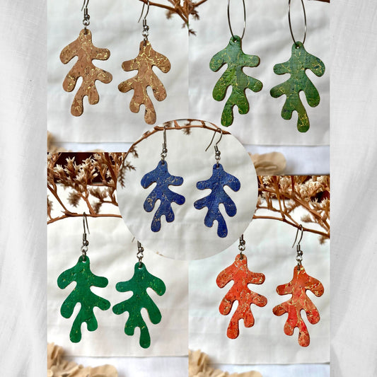 Five pairs of handpainted cork coral earrings in navy, emerald, orange, natural and khaki, displayed against a white linen background with dried botanicals.