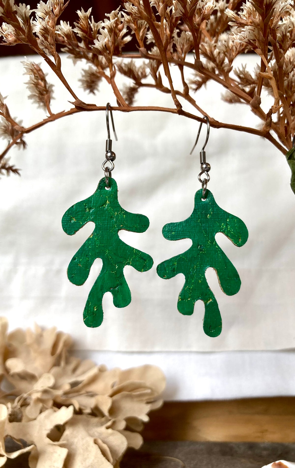 Emerald green cork coral earrings with gold fleck, hanging from a dried branch against a white background.