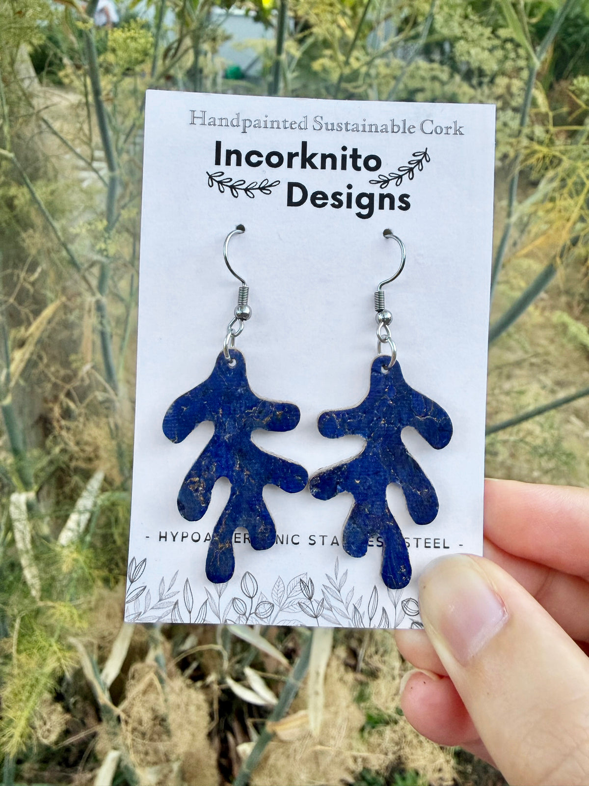 Navy blue handpainted cork coral earrings on an Incorknito Designs card, held outdoors against green foliage.