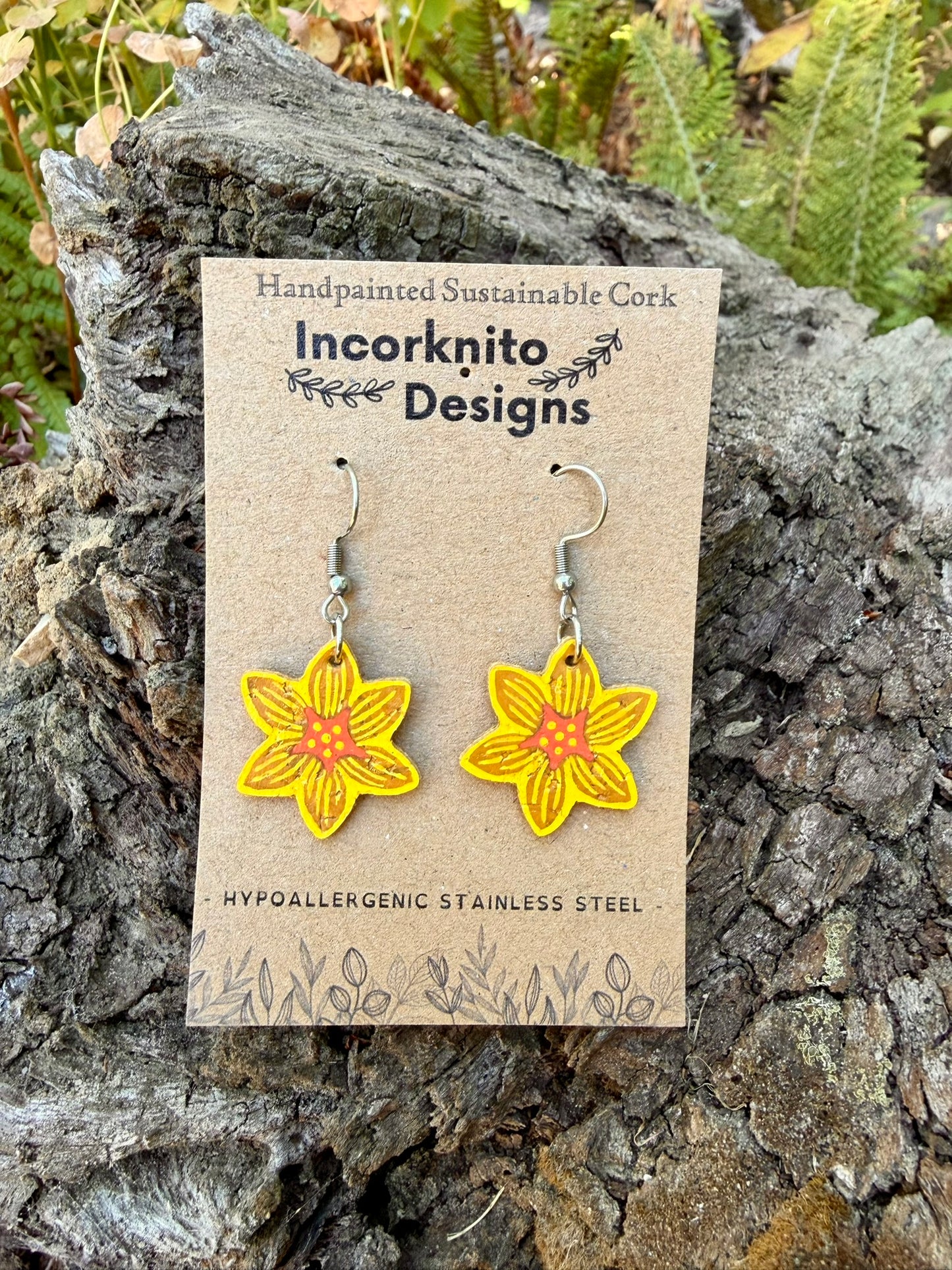 Cork daffodil hook earrings on an Incorknito Designs kraft card, propped against a mossy bark log with ferns in the background.