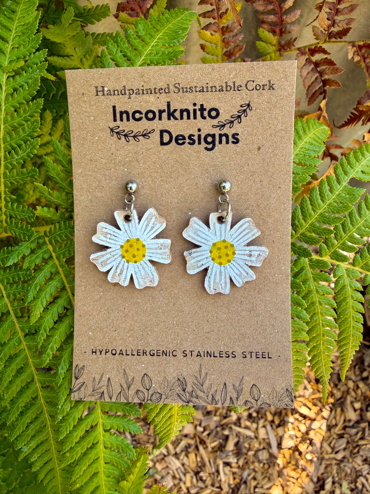 Handpainted cork daisy earrings on silver ball studs, displayed on Incorknito Designs branded card with fern background