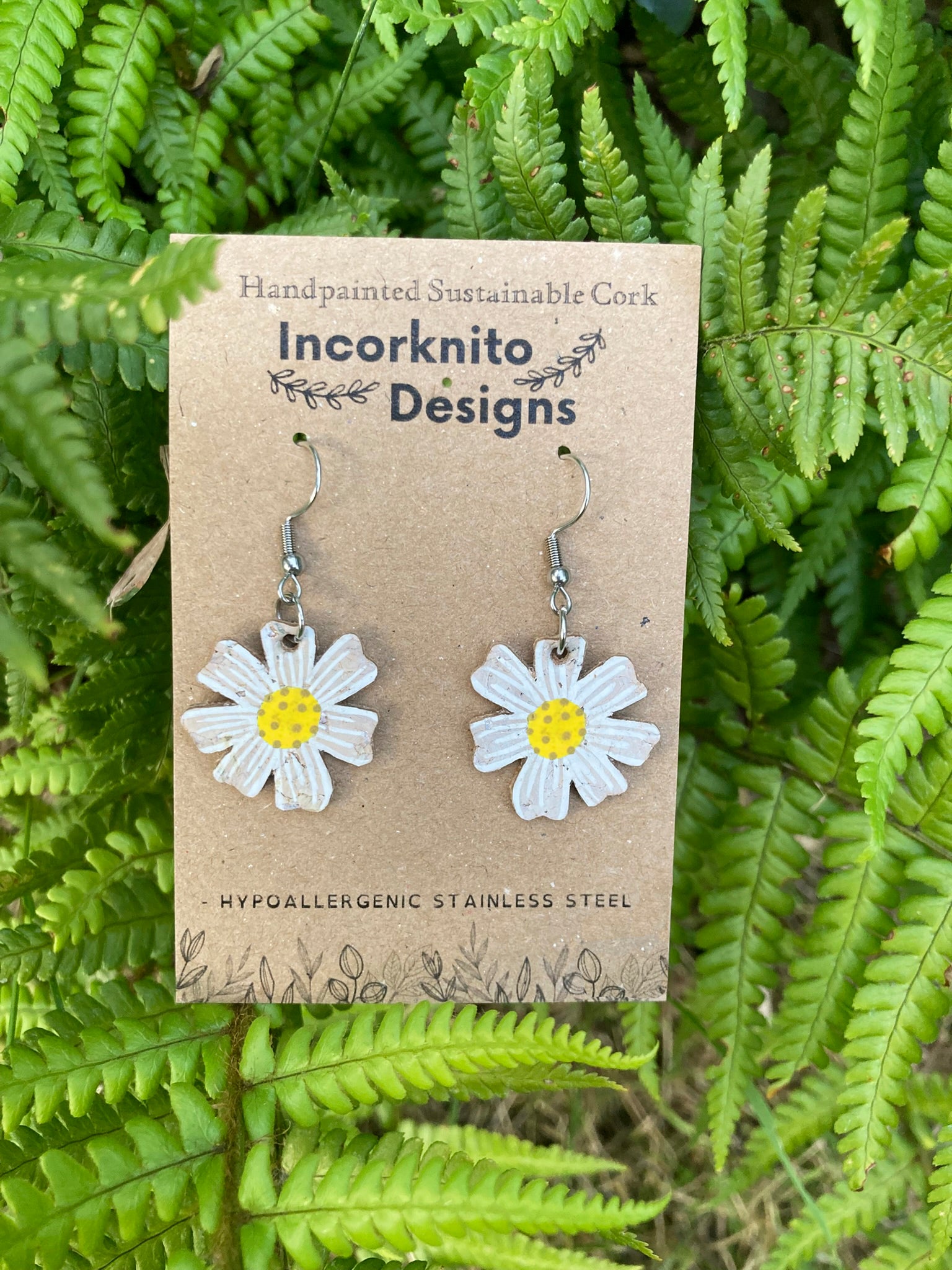 Handpainted cork daisy earrings on silver hooks, displayed on Incorknito Designs branded card with fern background