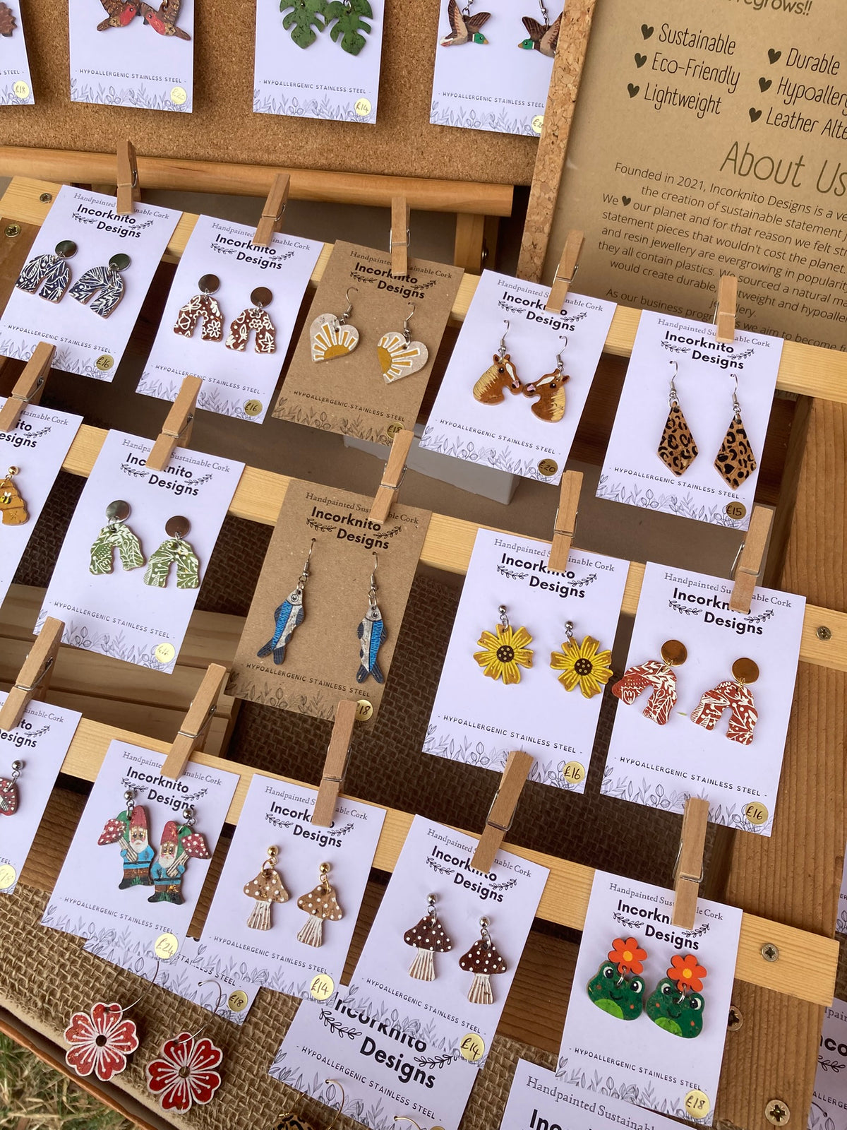 Detailed shot of a selection of Incorknito Designs handpainted cork earrings on branded backing cards attached to a wooden base with pegs. Designs include mushroom, gnome, sunflower and animal print styles