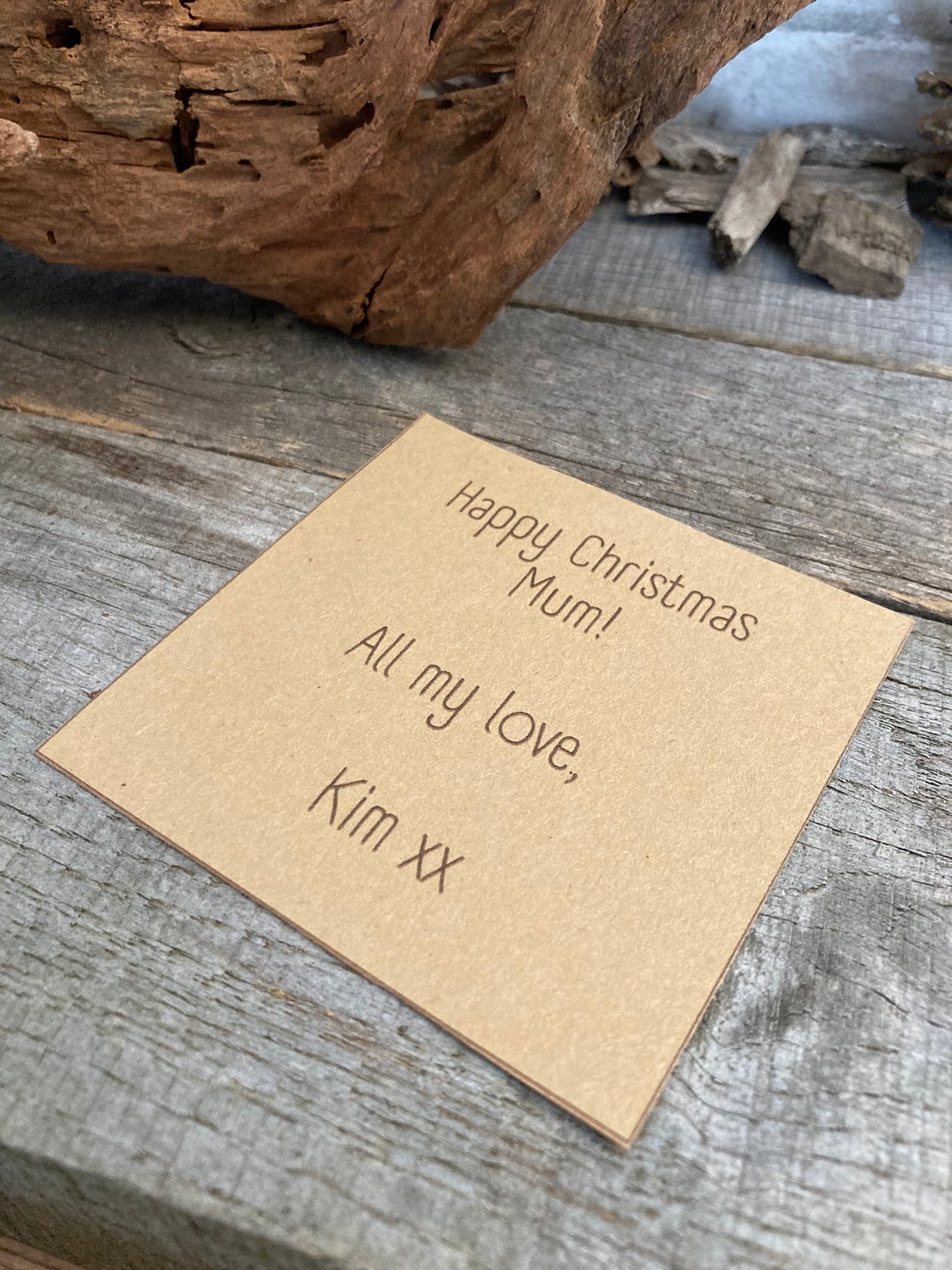 A personalised gift message card placed on a wooden surface, with the message 'Happy Christmas Mum! All my love, Kim xx' printed on it.