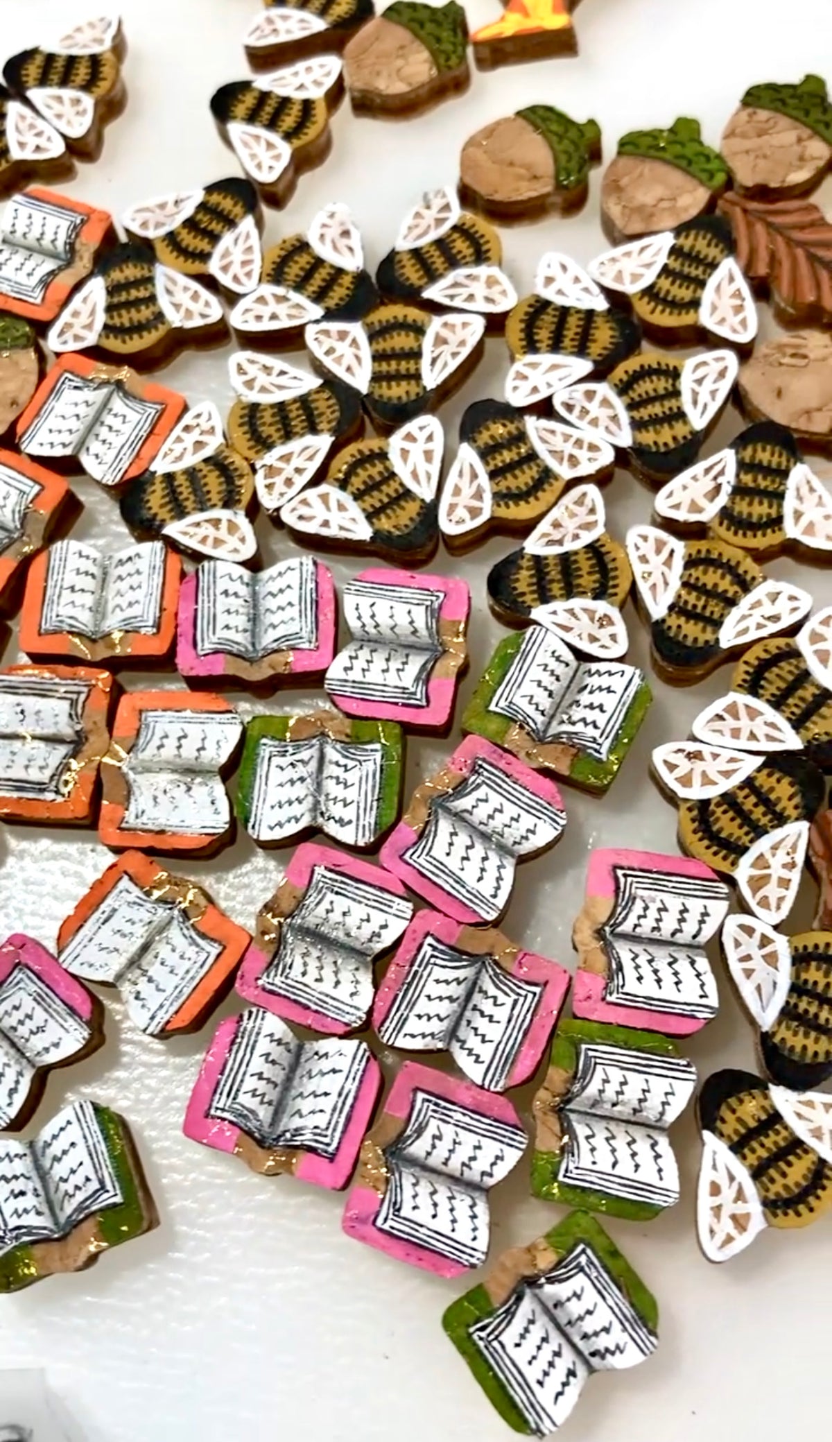 Dozens of hand-painted cork earrings laid out during production at Incorknito Designs, showing bee, acorn, leaf and open book designs at various stages of painting on a white surface.