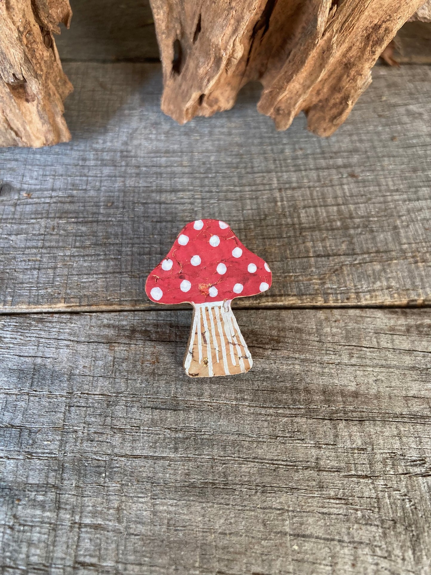 Red fly agaric toadstool cork pin badge handpainted with white spot detailing and gold fleck shimmer, handmade in Suffolk UK by Incorknito Designs resting on a flat lay of weathered wood.