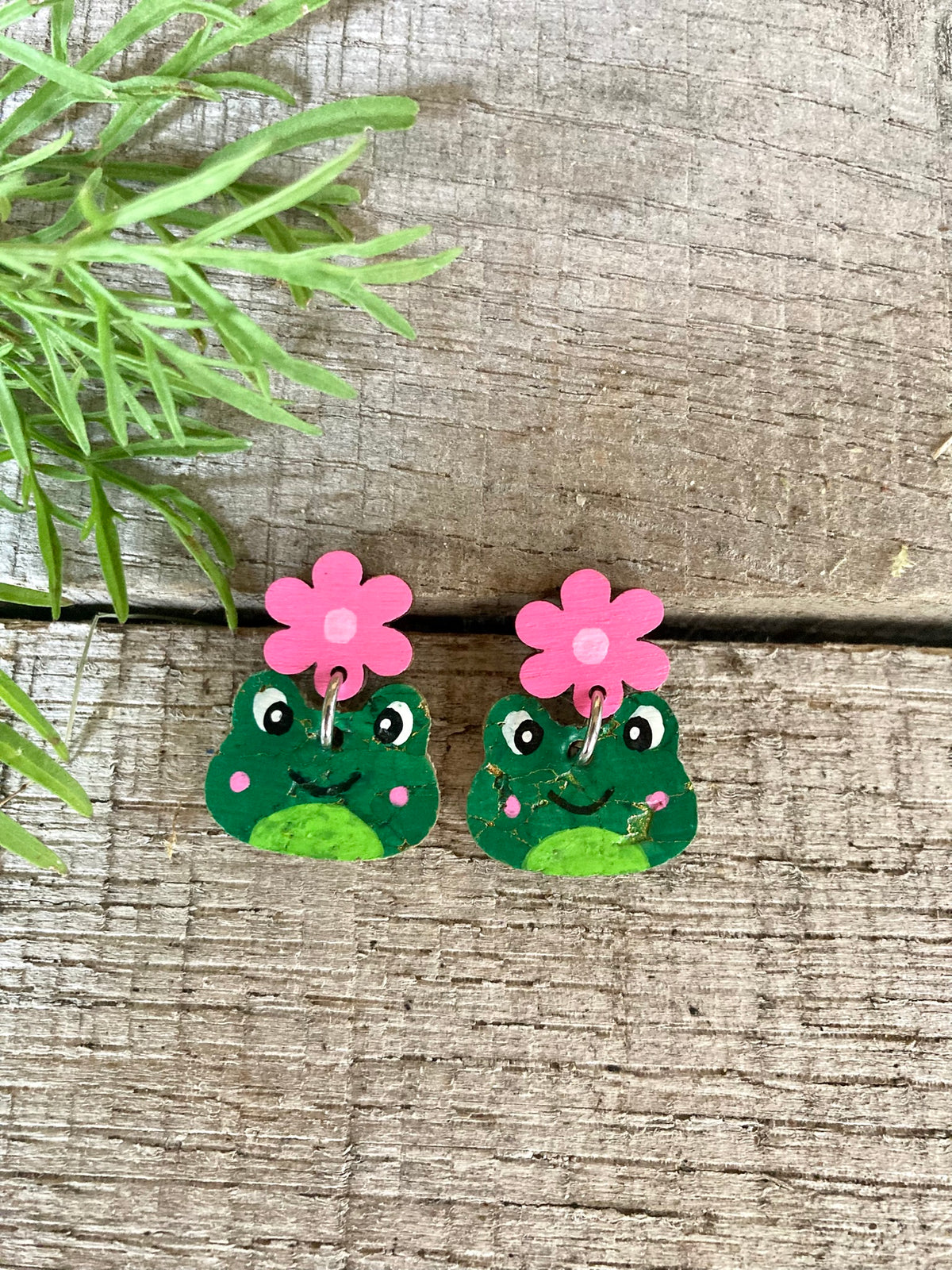 Handpainted cork frog flower stud drop earrings in pink, flat lay on weathered wood with green foliage