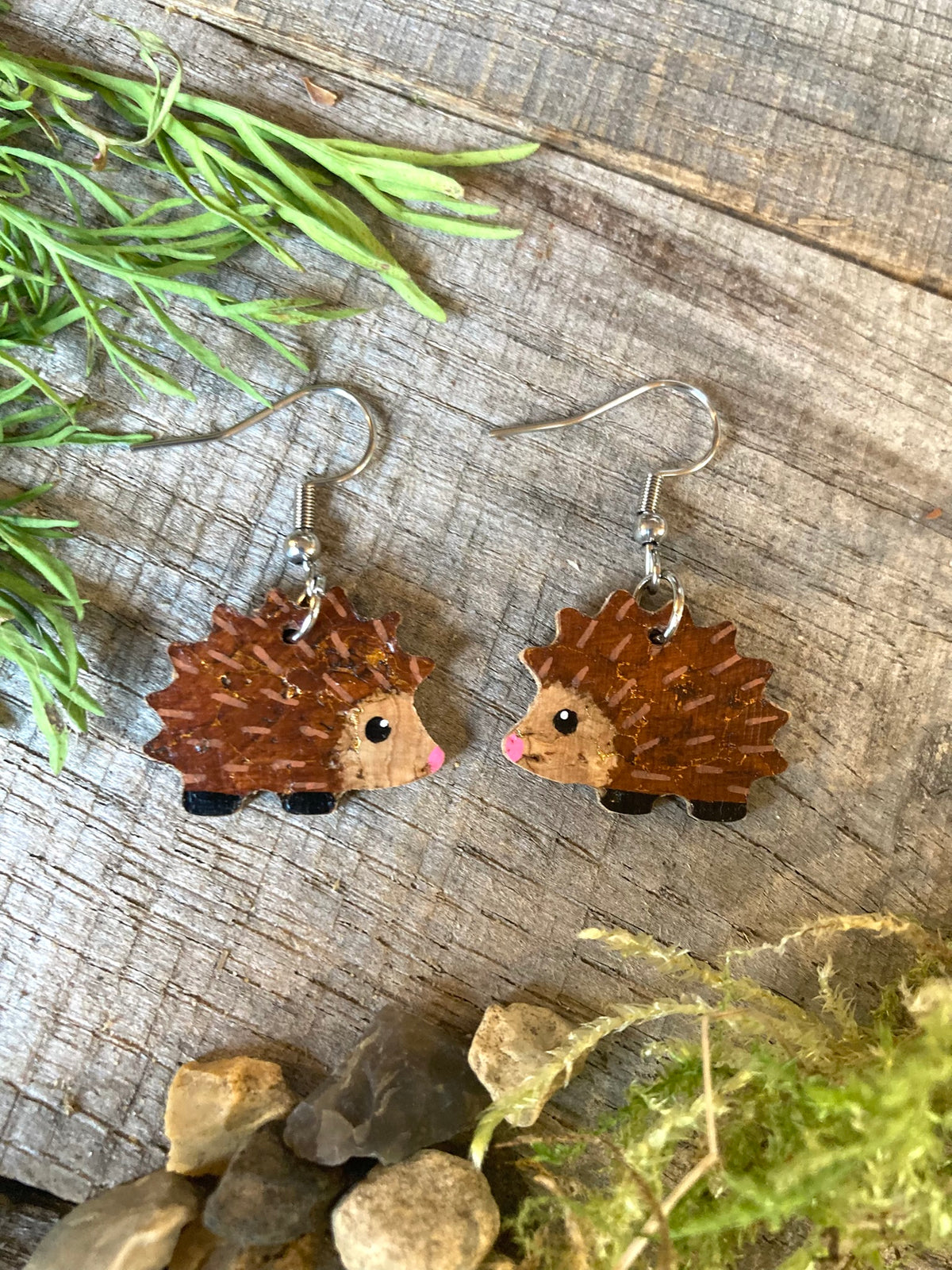 Handpainted hedgehog earrings on weathered wood surrounded by pine cones — natural cork face with gold fleck and brown spines