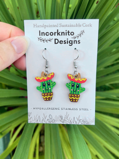 Handpainted cork cacti earrings with hook fastening on Incorknito Designs card, held against lush green palm leaves