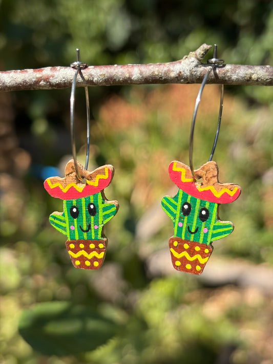 Handpainted cork Mexican cacti earrings on large silver hoops, hanging from a garden branch with blurred green foliage background
