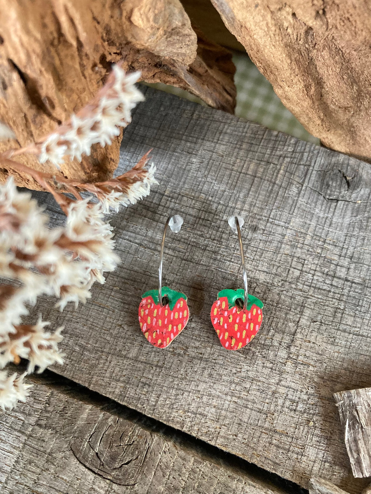 Handpainted mini strawberry cork earrings on silver open-back hoops, flat lay on weathered wood with dried white botanicals and cork bark