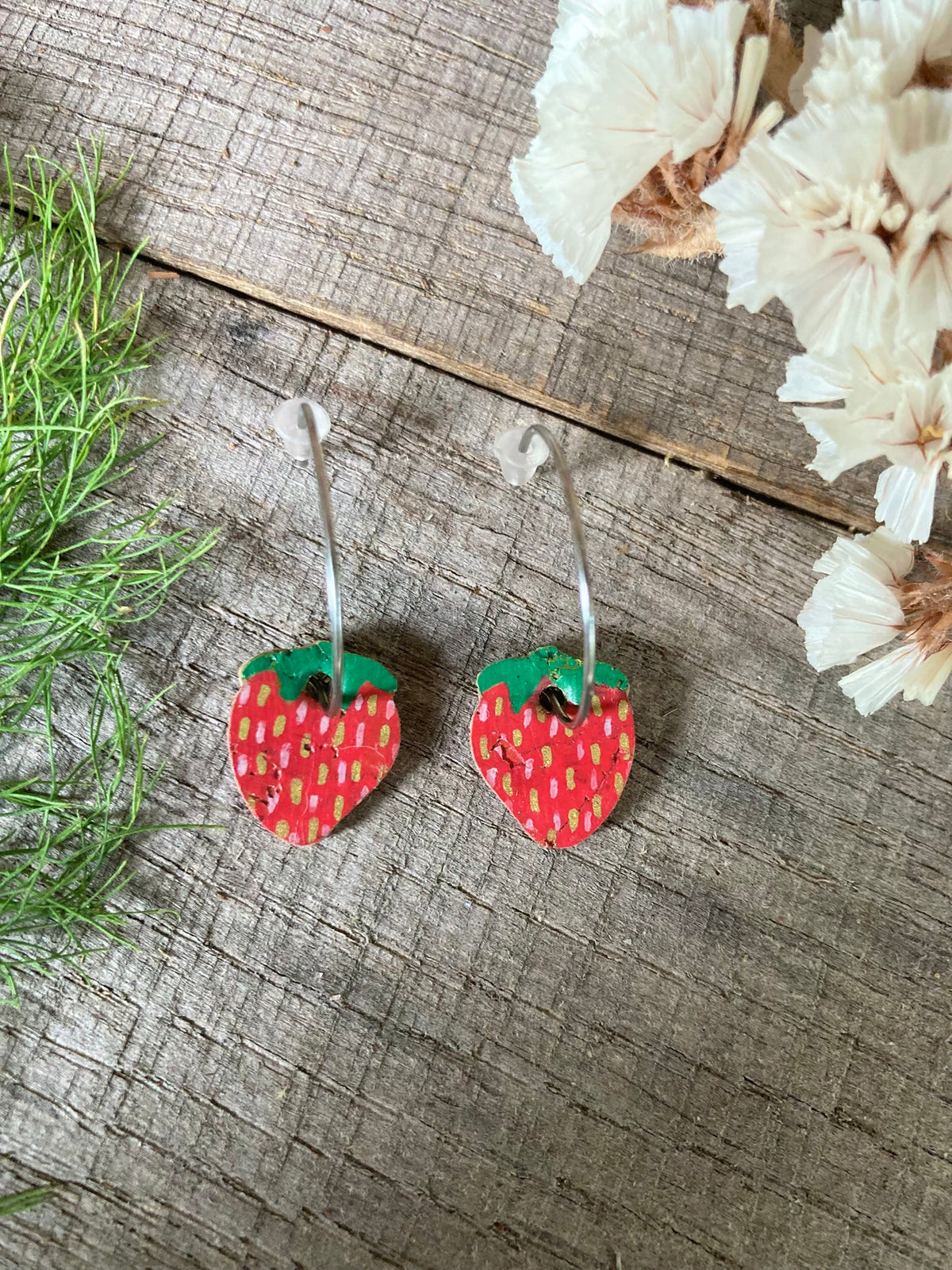 Handpainted mini strawberry cork earrings on silver open-back hoops, flat lay on weathered wood with green grass and dried white flowers