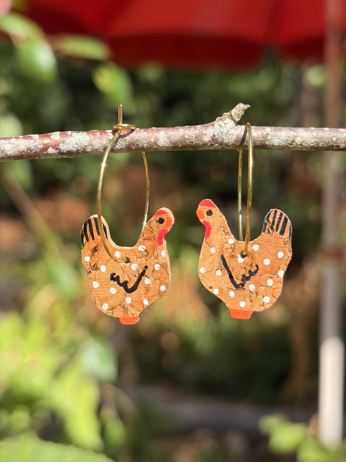Handpainted natural cork chicken earrings on gold hoops, hanging from a garden branch with soft green bokeh background