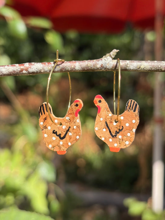 Handpainted natural cork chicken earrings on gold hoops, hanging from a garden branch with soft green bokeh background