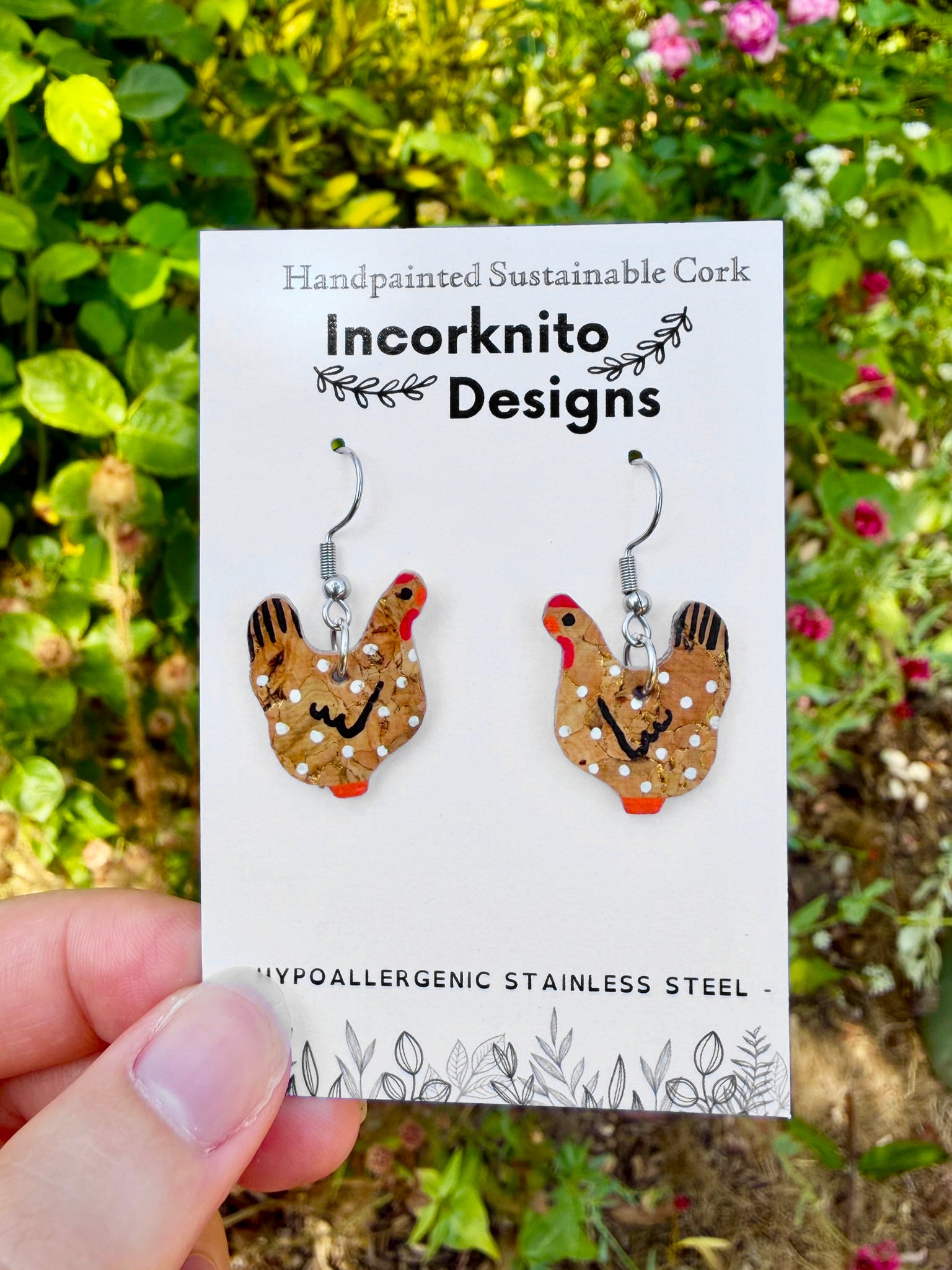 Handpainted natural cork chicken earrings on silver hooks, displayed on Incorknito Designs branded card held in hand against garden roses and greenery